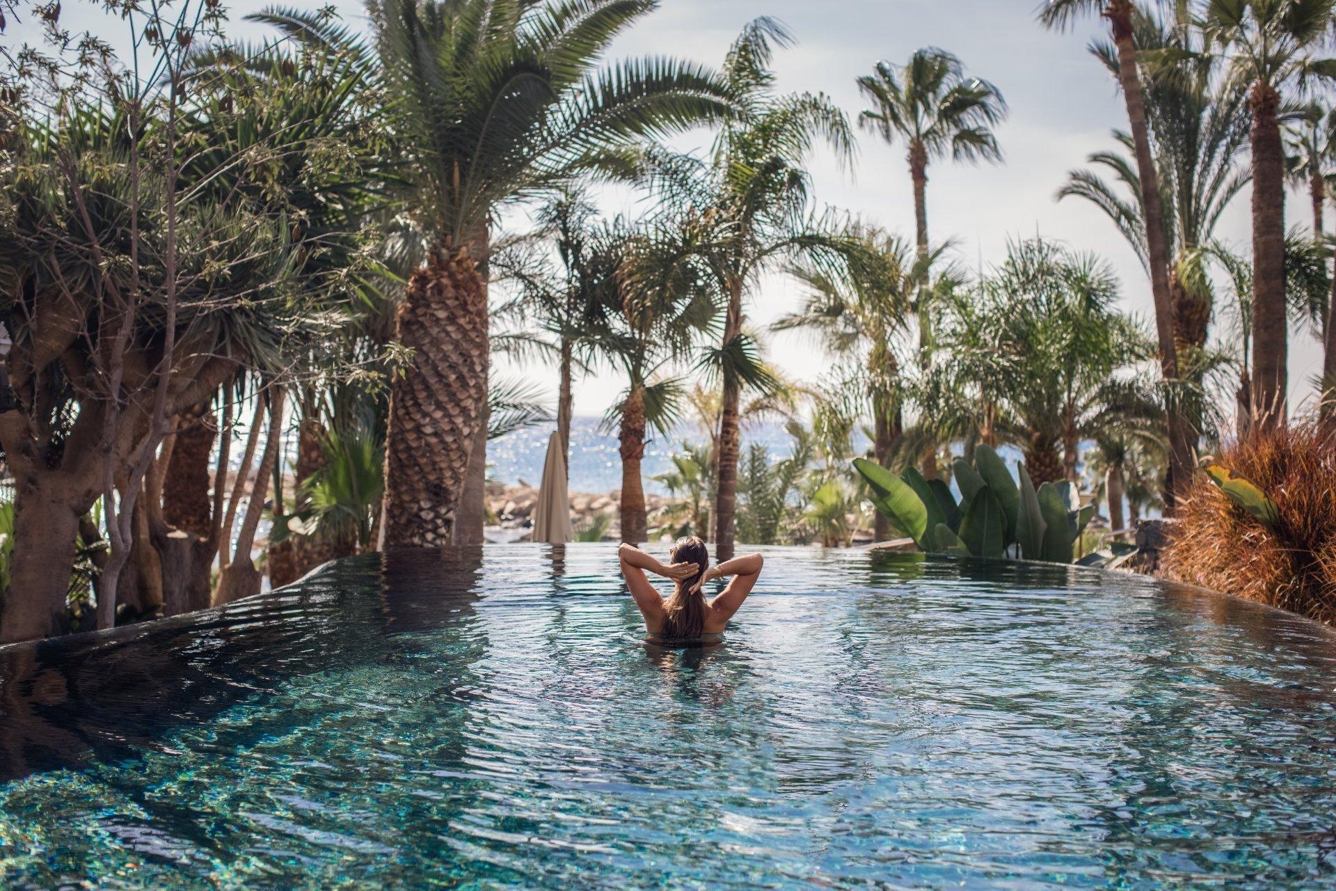mujer de espaldas en piscina tropical frente al mar rodeada de palmeras